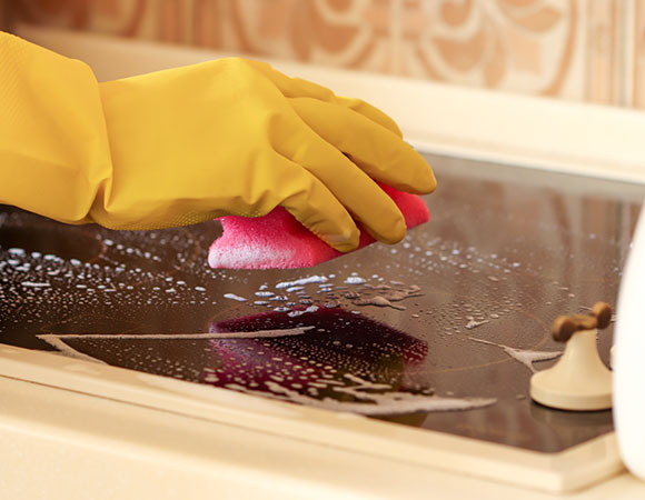 person wearing yellow rubber gloves cleaning a stovetop with a pink sponge and soap bubbles highlighting three cleaning steps for best results